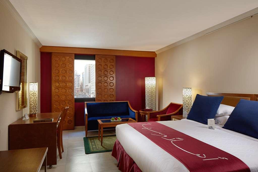 Makarem Ajyad Makkah Hotel Mecca Room photo The photo showcases a hotel room featuring a modern design. In the center, there's a large bed with white linens and a decorative red runner. On either side of the bed, there are wooden nightstands with lamps. A blue sitting area is visible, composed